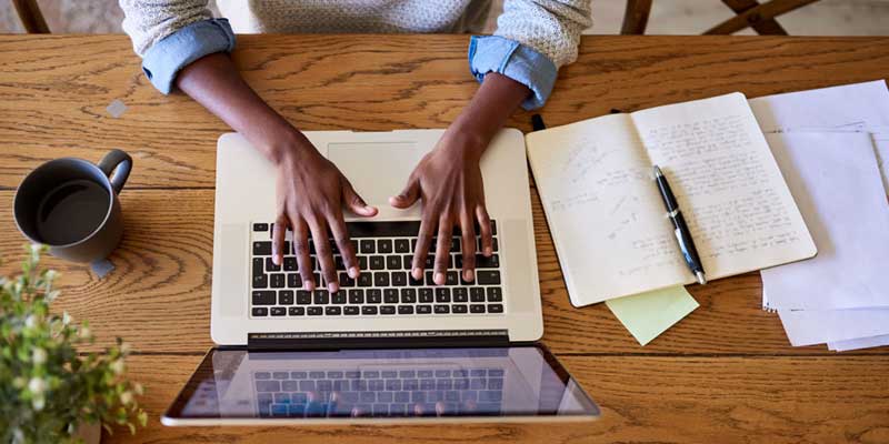 Image: An overhead shot of an adult's hands on the keyboard, with a coffee mug and various papers/notebooks open on the desk. Image: An overhead shot of an adult's hands on the keyboard, with a coffee mug and various papers/notebooks open on the desk.